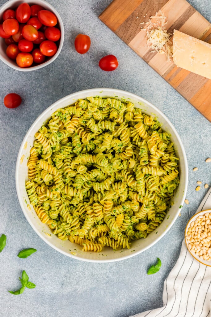 A large white bowl of Pesto Caprese Pasta Salad with rotini coated in green pesto sauce sits on a gray surface, surrounded by cherry tomatoes, a block of Parmesan cheese on a wooden board, pine nuts, and fresh basil leaves.