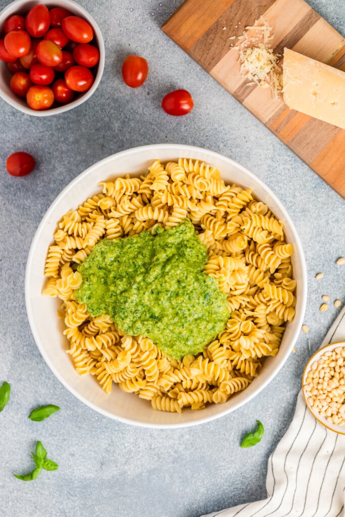 A bowl of rotini pasta topped with green pesto sauce sits on a gray surface, surrounded by cherry tomatoes, Parmesan cheese, pine nuts, and fresh basil—perfect for making a vibrant Pesto Caprese Pasta Salad.