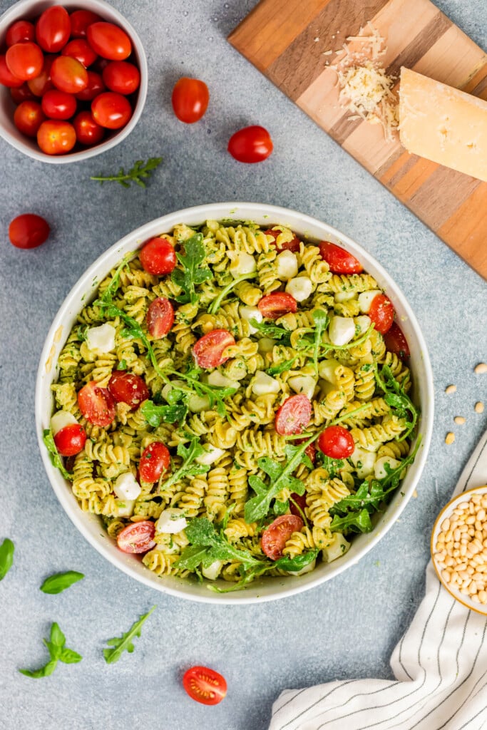 A bowl of Pesto Caprese Pasta Salad with cherry tomatoes, mozzarella balls, and arugula. Nearby are a bowl of cherry tomatoes, a wedge of Parmesan on a wooden board, pine nuts, and a striped cloth.