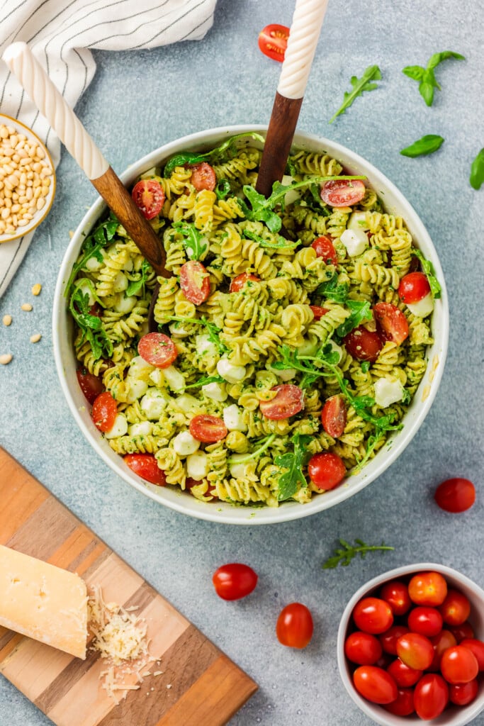 A bowl of Pesto Caprese Pasta Salad with green pesto, cherry tomatoes, mozzarella balls, and arugula is served with wooden utensils. Around the bowl are pine nuts, Parmesan cheese, extra cherry tomatoes, and a striped napkin.