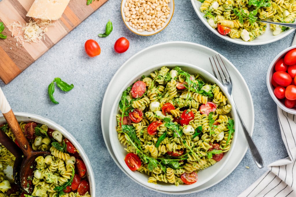 A bowl of Pesto Caprese Pasta Salad with cherry tomatoes, mozzarella, and arugula sits on a table. Nearby are more salad bowls, a cutting board with cheese, a small bowl of pine nuts, and fresh basil leaves.