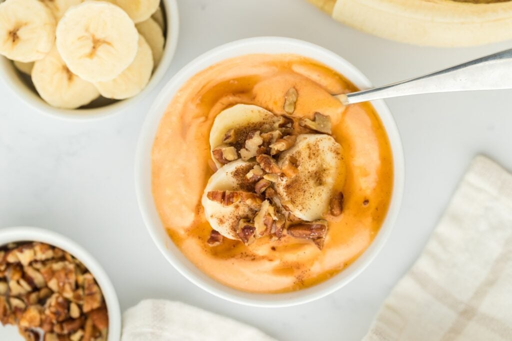 A bowl of creamy orange smoothie, perfect for high protein snacks, topped with banana slices, chopped nuts, and a sprinkle of cinnamon. Sliced bananas and nuts are in small bowls nearby on a white surface, with a spoon in the bowl.