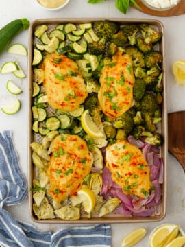 A sheet pan with four pieces of baked, cheesy Roasted Garlic Hummus Chicken & Veggies (Easy One Sheet Dinner) surrounded by roasted zucchini, broccoli, red onion, and artichoke hearts. Lemon wedges, hummus, fresh herbs, and a blue-striped towel are nearby.
