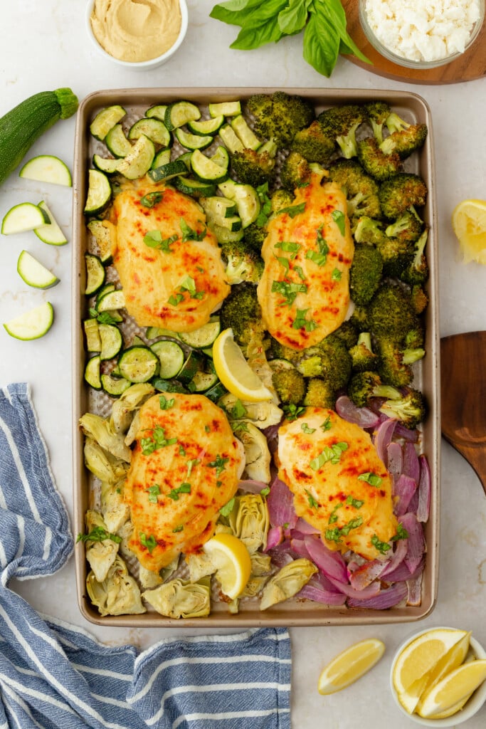 A sheet pan with four pieces of baked, cheesy Roasted Garlic Hummus Chicken & Veggies (Easy One Sheet Dinner) surrounded by roasted zucchini, broccoli, red onion, and artichoke hearts. Lemon wedges, hummus, fresh herbs, and a blue-striped towel are nearby.