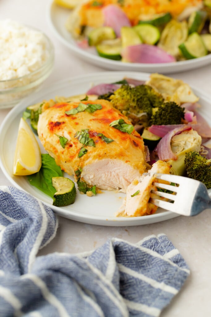 A plate of Roasted Garlic Hummus Chicken & Veggies (Easy One Sheet Dinner) is garnished with herbs and served with roasted broccoli, zucchini, red onions, and a lemon wedge. A fork holds a bite, while a blue-striped napkin and small bowl of cheese sit nearby.