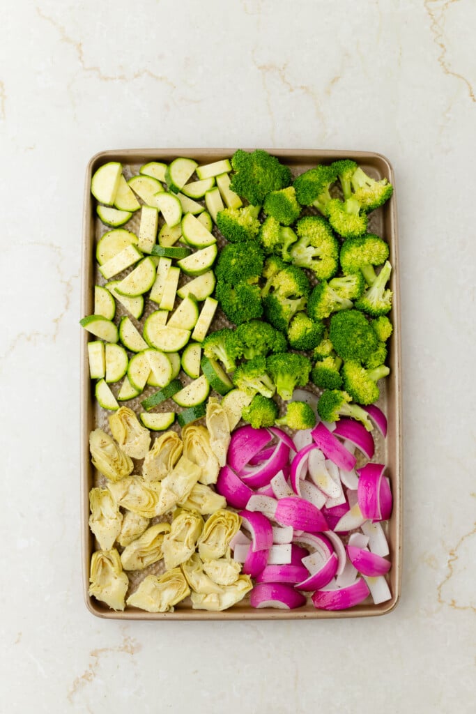 A baking sheet with chopped zucchini, broccoli florets, red onion slices, and artichoke hearts arranged in separate sections—perfect for preparing Roasted Garlic Hummus Chicken & Veggies (Easy One Sheet Dinner) on a light-colored countertop.