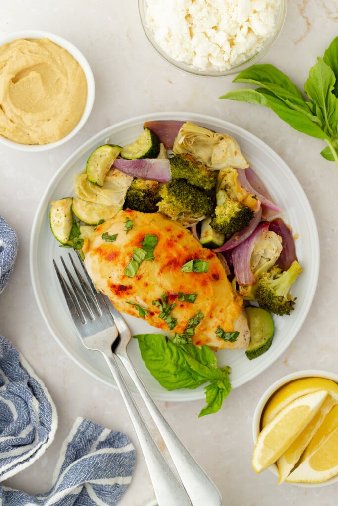 A white plate with Roasted Garlic Hummus Chicken & Veggies (Easy One Sheet Dinner) features baked chicken breast topped with herbs, surrounded by roasted broccoli, zucchini, artichoke, and red onion. Lemon wedges, basil, feta cheese, and a blue napkin complete the scene.