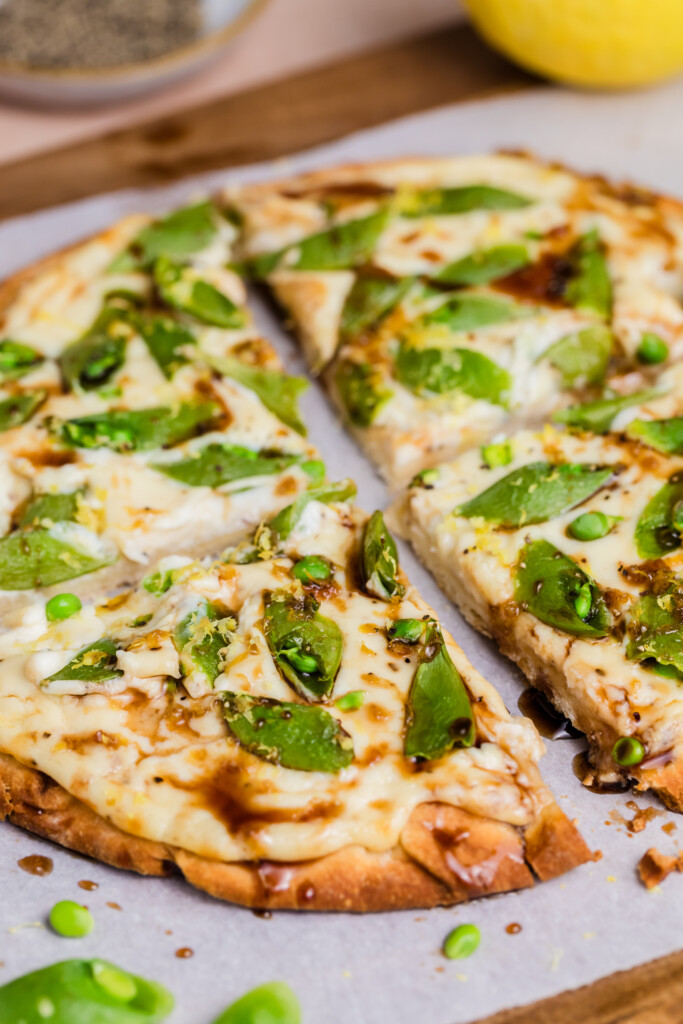 A sliced flatbread pizza topped with melted cheese, sugar snap peas, and a drizzle of sauce, displayed on parchment paper atop a wooden board.