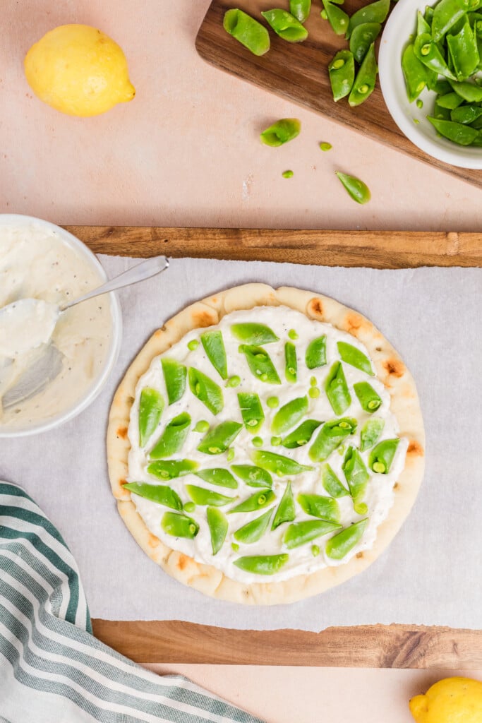 A flatbread topped with a creamy white spread and sliced snap peas sits on parchment paper. Nearby are a bowl of snap peas, a lemon, a green-striped cloth, and a bowl of white sauce with a spoon.