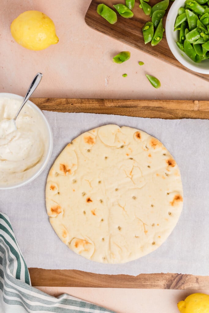 A round flatbread sits on parchment paper with a bowl of white sauce and a spoon to the left, sliced snap peas in a bowl above, lemons, and a green-striped cloth in the corner.