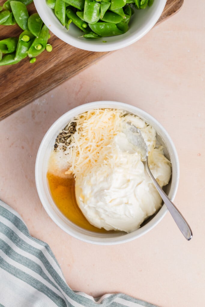 A white bowl with sour cream, shredded cheese, seasonings, and a spoon sits on a light surface near a striped cloth; a cutting board with cut green beans and a bowl of green beans are nearby.