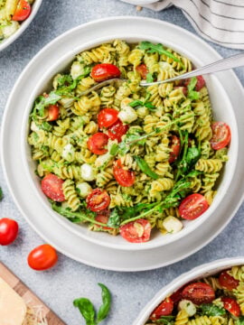 A bowl of Pesto Caprese Pasta Salad with rotini, cherry tomatoes, arugula, and mozzarella balls, surrounded by extra salad bowls, fresh tomatoes, basil leaves, and a striped napkin on a light surface.