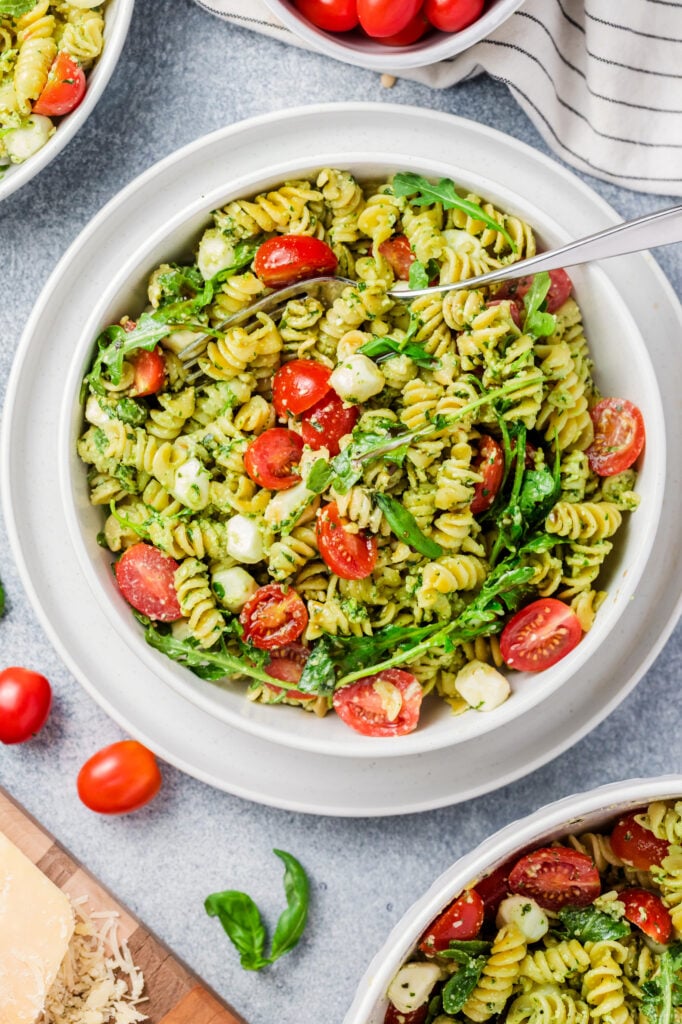A bowl of Pesto Caprese Pasta Salad with rotini, cherry tomatoes, arugula, and mozzarella balls, surrounded by extra salad bowls, fresh tomatoes, basil leaves, and a striped napkin on a light surface.