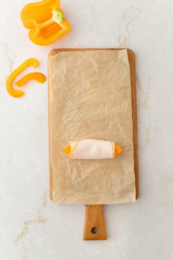 A wooden cutting board lined with parchment paper holds a piece of yellow bell pepper wrapped in turkey. Slices of yellow bell pepper and a pepper top are on the white countertop beside the board.