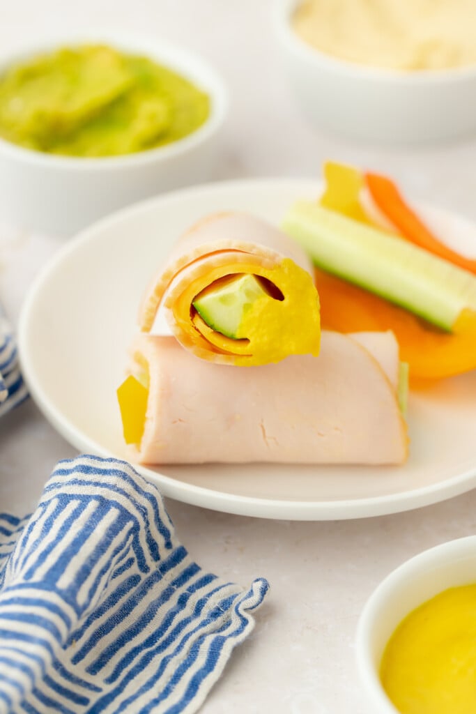 A plate with turkey slices wrapped around cucumber and yellow bell pepper, surrounded by small bowls of green dip, yellow sauce, and sliced vegetables, with a blue and white striped napkin nearby.