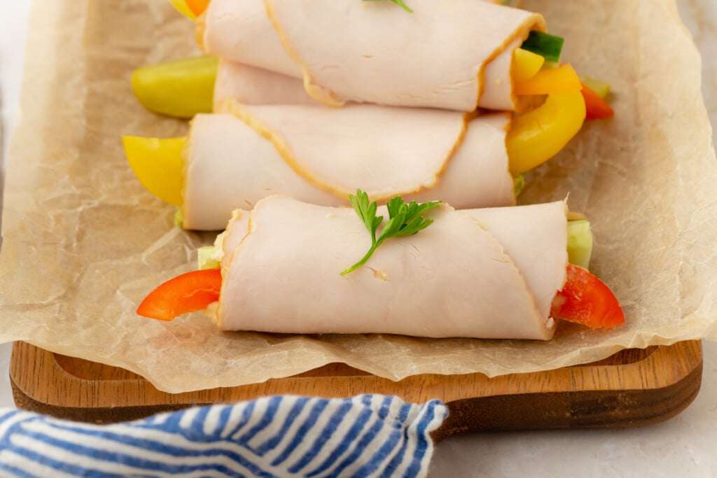Three turkey roll-ups filled with sliced red and yellow bell peppers and cucumber are placed on parchment paper. A parsley leaf garnishes the front roll-up. A wooden board and blue striped cloth are visible underneath.