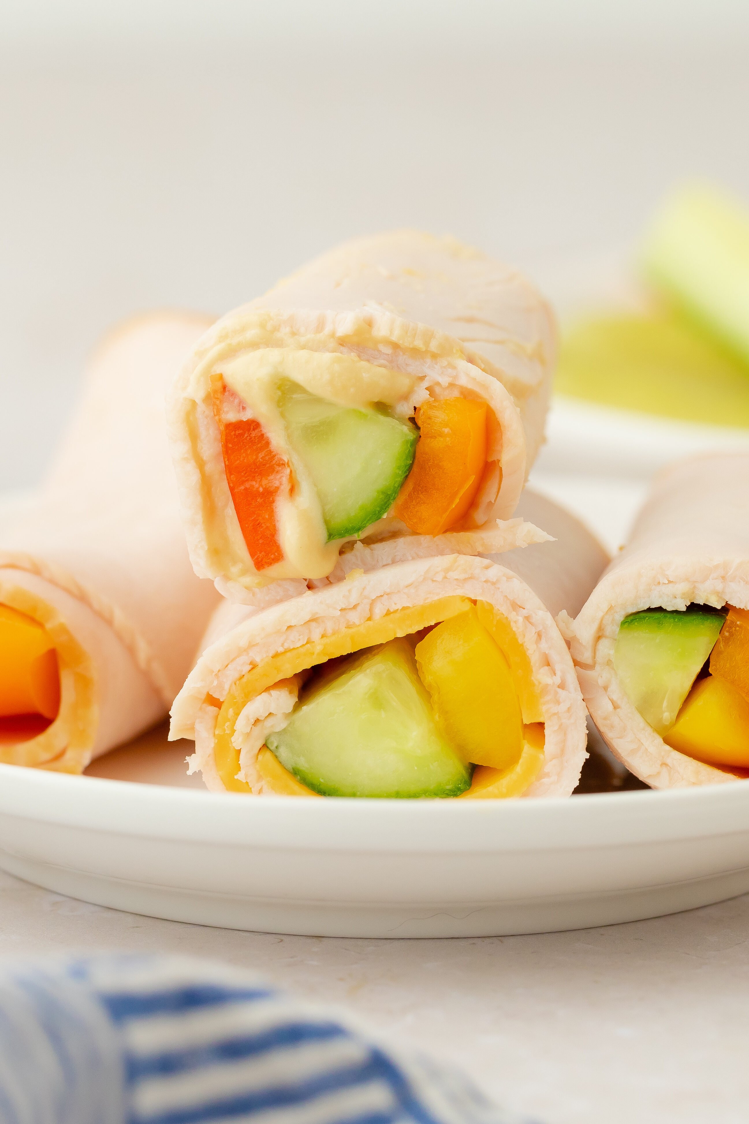 Four turkey roll-ups filled with sliced cucumber, bell pepper, and carrots are arranged on a white plate. In the background are bowls with guacamole and more sliced vegetables. A blue striped cloth is in the foreground.