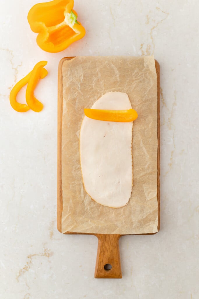 A slice of deli turkey breast on parchment paper atop a wooden cutting board, with a strip of yellow bell pepper placed on the turkey. Nearby, there’s a cut yellow bell pepper and another pepper slice on a light surface.