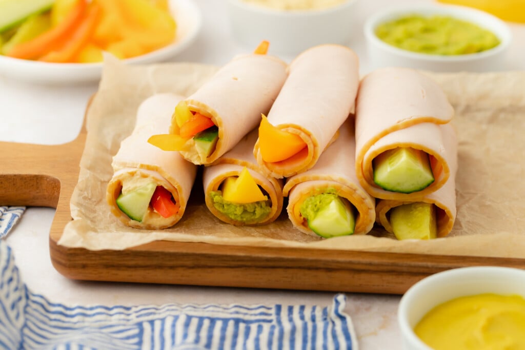 Six turkey roll-ups filled with sliced vegetables and avocado are arranged on parchment paper atop a wooden tray. Small bowls of dip and more vegetable slices are visible in the background.