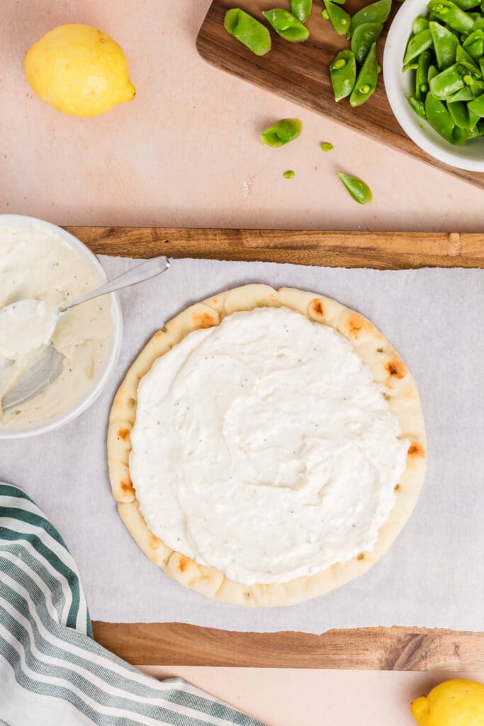 Flatbread pizza base being topped with a creamy white spread from a bowl, with sugar snap peas and a lemon nearby.
