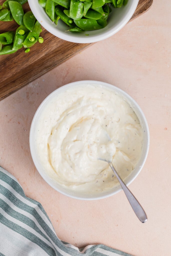 A bowl of creamy white dip with a spoon in it, placed on a light surface near a striped cloth and a cutting board with chopped green vegetables.