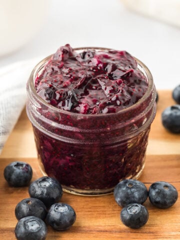 A small glass jar filled with thick, dark purple blueberry jam sits on a wooden surface, surrounded by fresh blueberries. A white cloth is partially visible in the background.