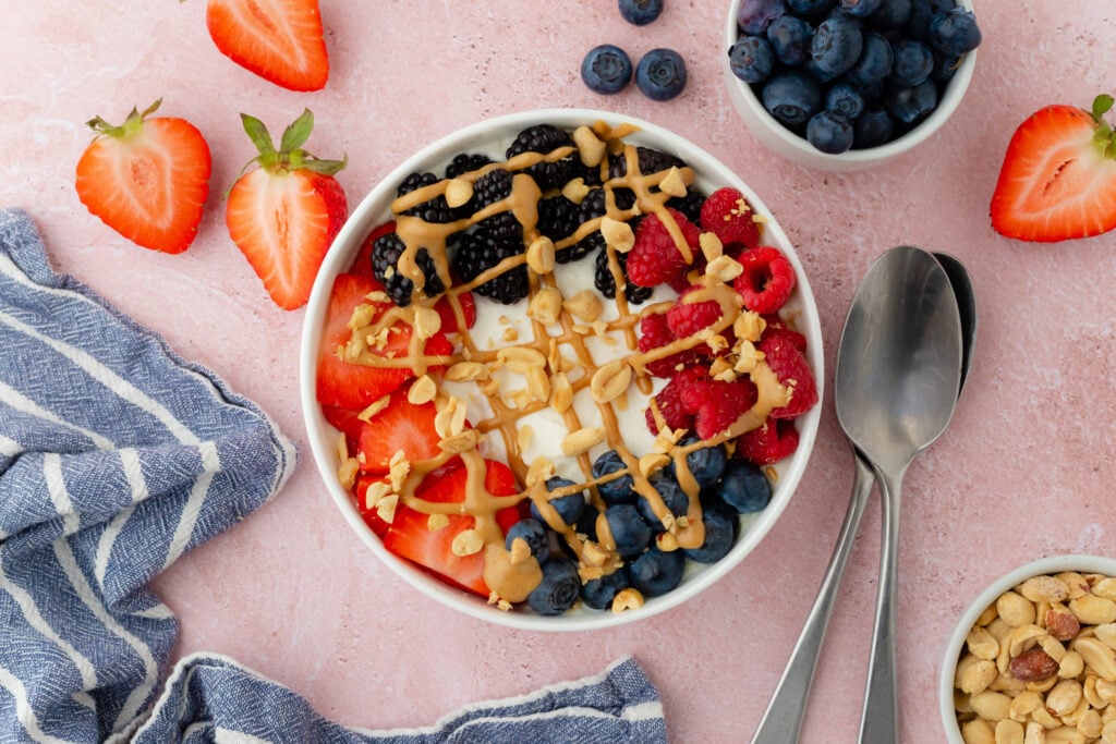 A bowl of yogurt with strawberries, blackberries, raspberries, blueberries, chopped nuts, and a drizzle of nut butter – a delicious option for those looking for high-fiber breakfast recipes. Bowls of chopped strawberries and nuts add extra flavor to the pink surface.