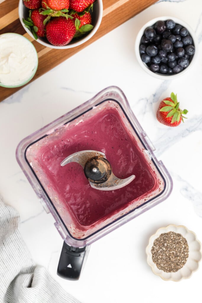 A blender filled with a thick, purple smoothie sits on a countertop surrounded by fresh strawberries, blueberries, a bowl of chia seeds, and a dish of yogurt.