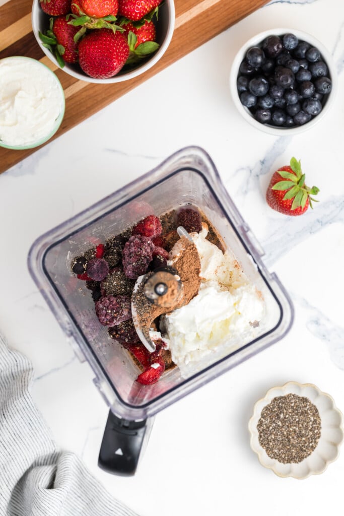 Top-down view of a blender filled with frozen berries, yogurt, nut butter, chia seeds, and other smoothie ingredients. Surrounding the blender are bowls of strawberries, blueberries, chia seeds, and a towel.