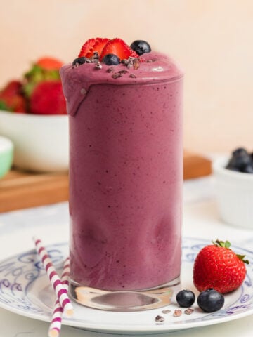 A tall glass of berry smoothie topped with sliced strawberries and blueberries sits on a plate with striped straws. Fresh strawberries and blueberries are scattered around, with bowls of fruit in the background.