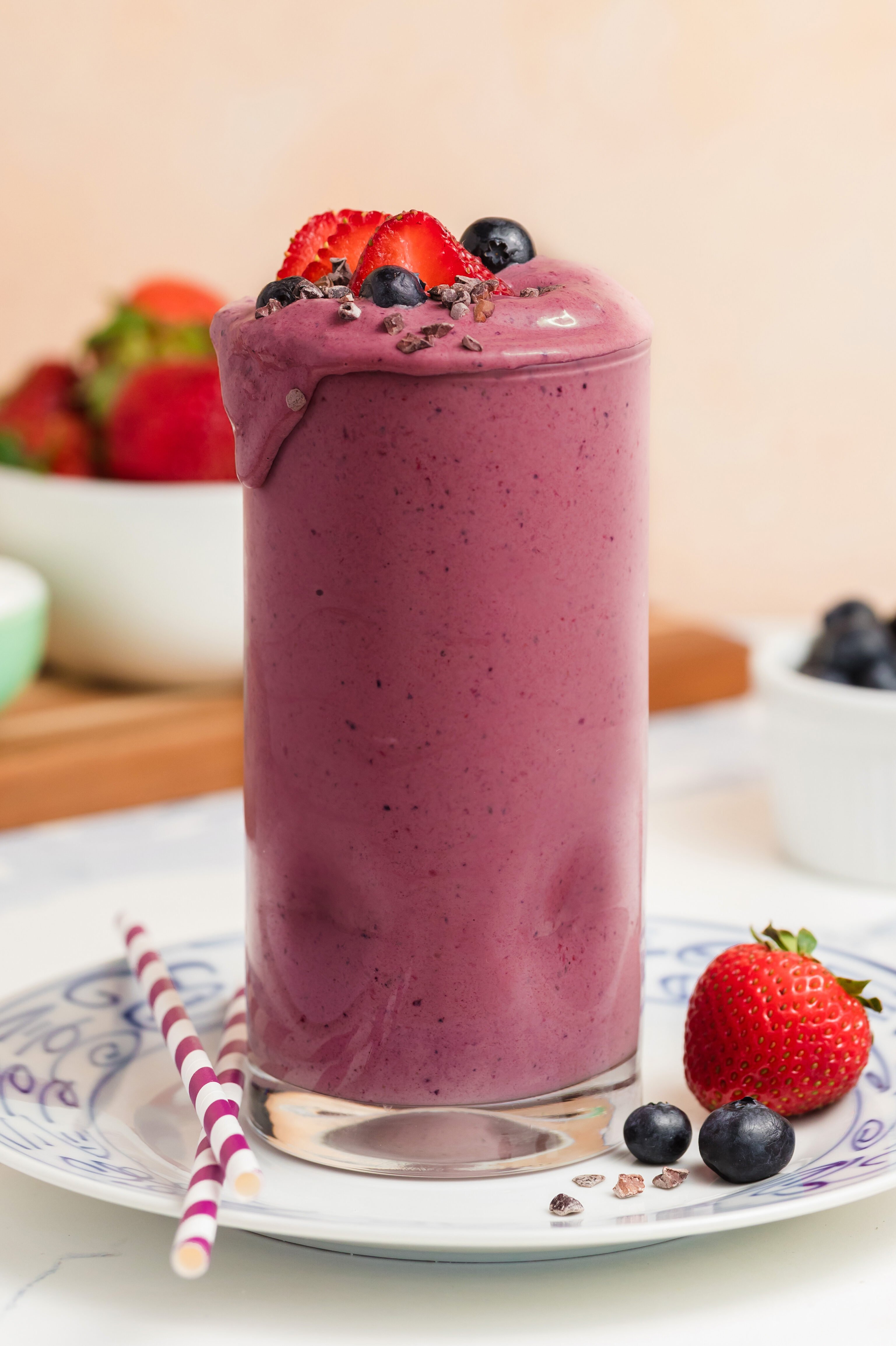A tall glass of berry smoothie topped with sliced strawberries and blueberries sits on a plate with striped straws. Fresh strawberries and blueberries are scattered around, with bowls of fruit in the background.