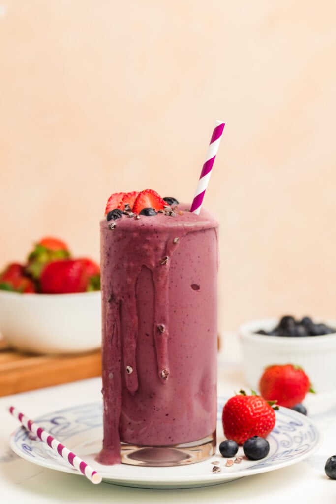 A tall glass of purple berry smoothie with a striped straw, topped with fresh strawberries and blueberries. Smoothie is overflowing, and whole berries are scattered around, with bowls of fruit in the background.