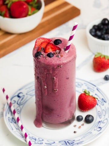 A tall glass of purple berry smoothie topped with fresh blueberries and sliced strawberries, with a striped straw. Surrounded by strawberries, blueberries, and ingredients on a white surface.