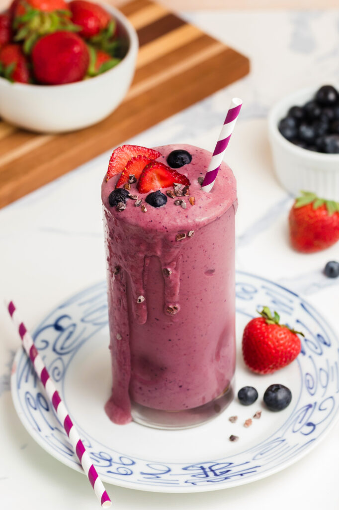 A tall glass of purple berry smoothie topped with fresh blueberries and sliced strawberries, with a striped straw. Surrounded by strawberries, blueberries, and ingredients on a white surface.