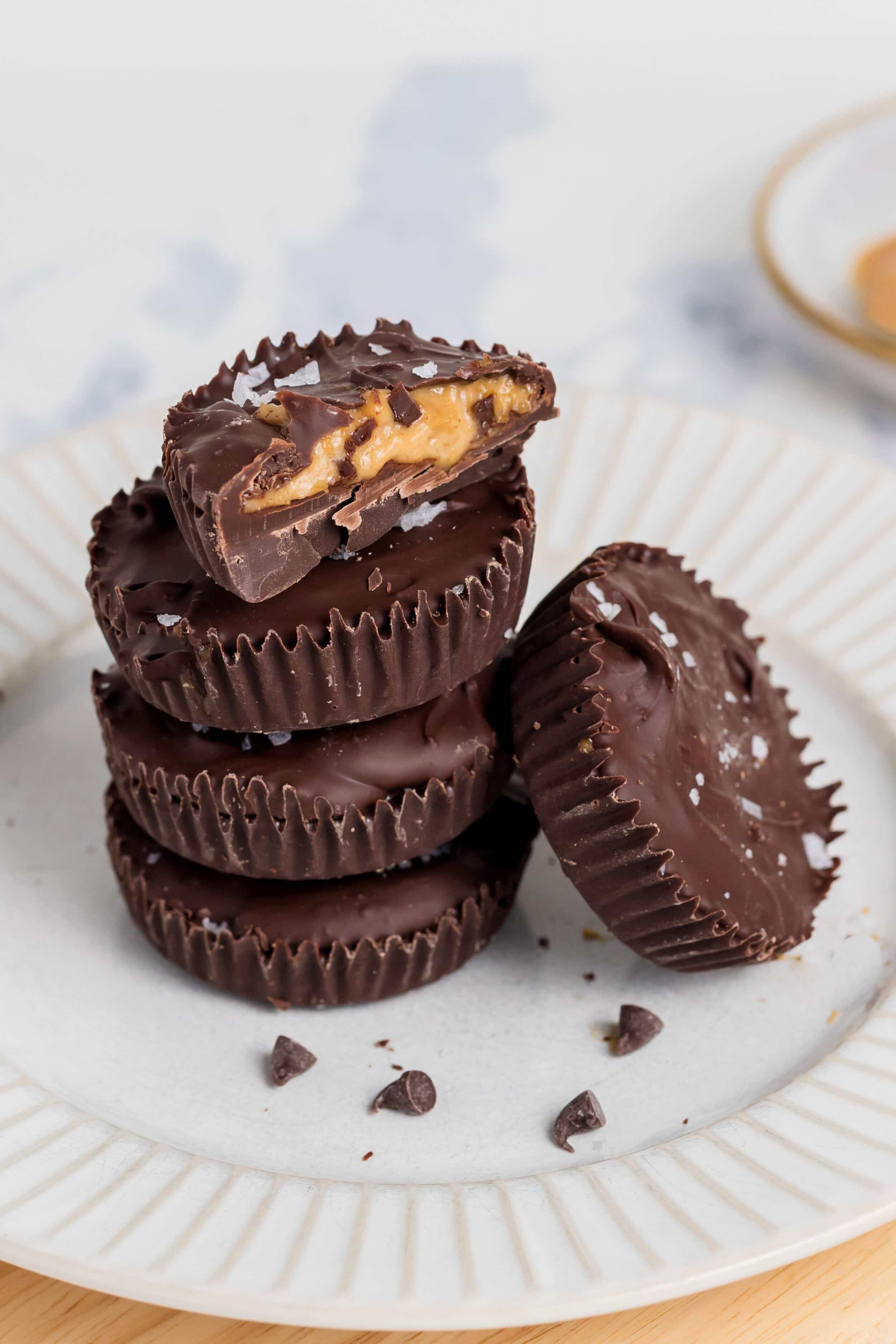 A stack of Chocolate-PB Cups sits on a white plate, with the top cup cut in half to reveal the creamy peanut butter filling. Other cups and a bowl of peanut butter are visible in the background.