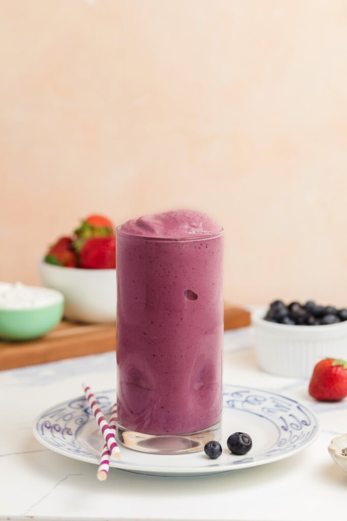 A tall glass filled with a thick purple smoothie sits on a patterned plate with two striped straws. Blueberries, strawberries, and bowls of ingredients are in the background.