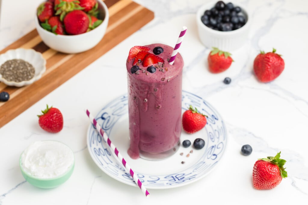 A tall glass of purple berry smoothie topped with strawberries, blueberries, and chia seeds sits on a white and blue plate, surrounded by fresh berries, chia seeds, and a bowl of yogurt on a marble surface.