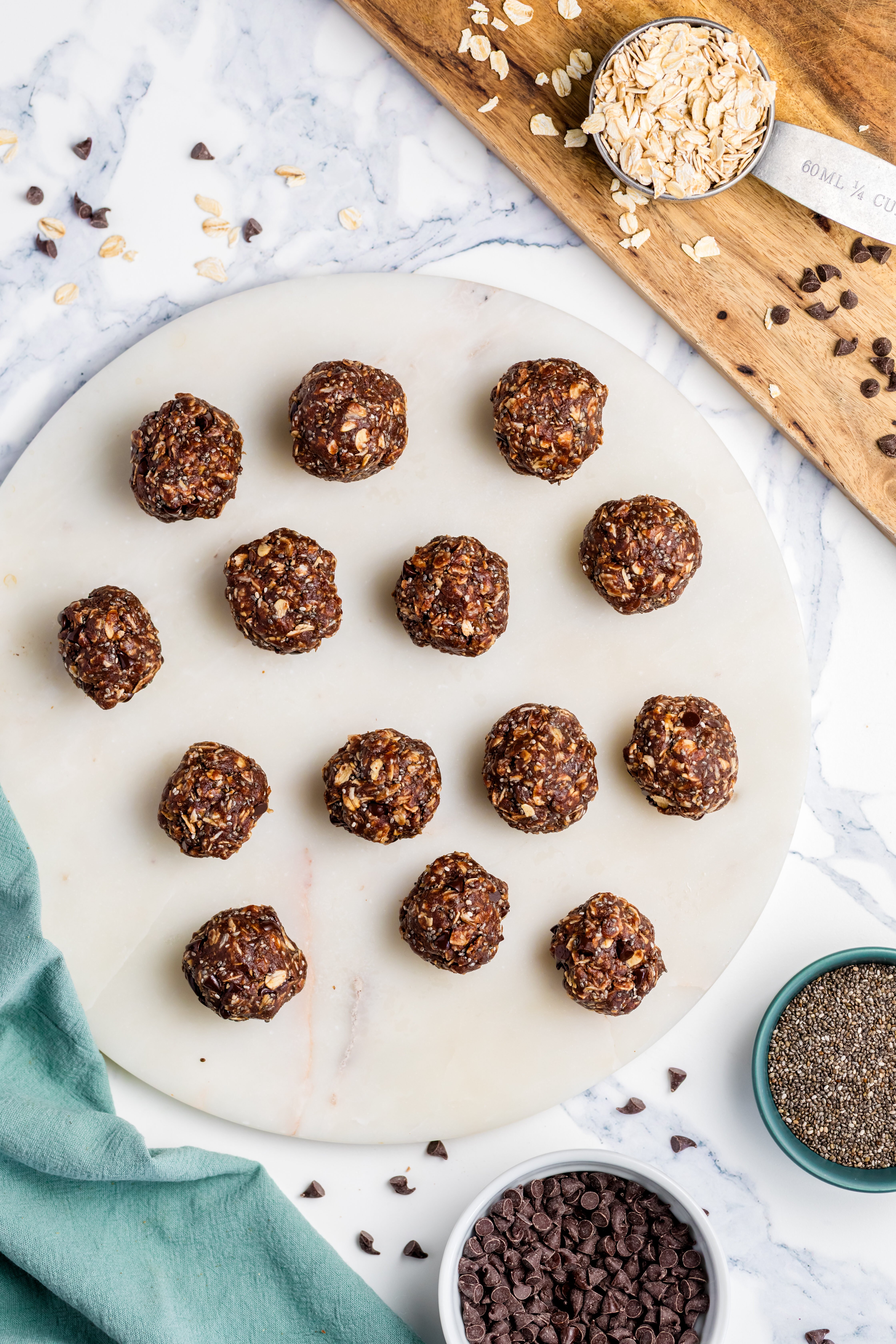 A marble platter with 15 Chocolate Peanut Butter Energy Bites, surrounded by bowls of chia seeds, chocolate chips, oats, and a green napkin on a white marble surface.