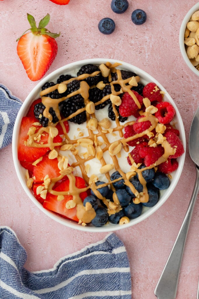 A PB and Berry Breakfast Bowl featuring yogurt topped with strawberries, blackberries, raspberries, blueberries, granola, and a drizzle of peanut butter sits on a pink surface beside a striped napkin, two spoons, nuts, and extra berries.