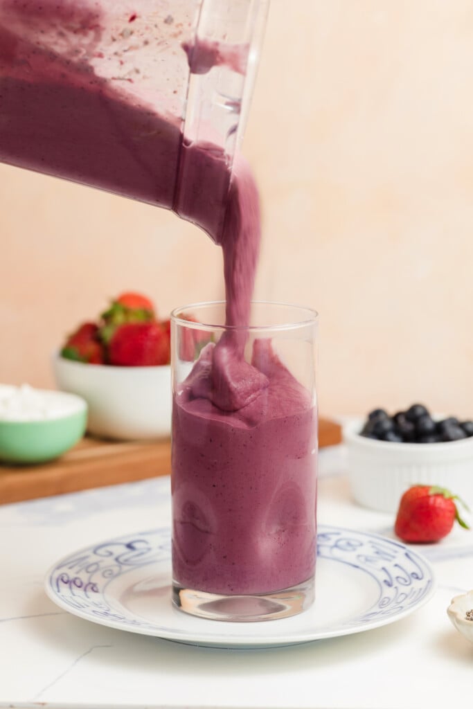 A purple smoothie is poured from a blender into a glass on a plate, with strawberries and blueberries in bowls in the background.