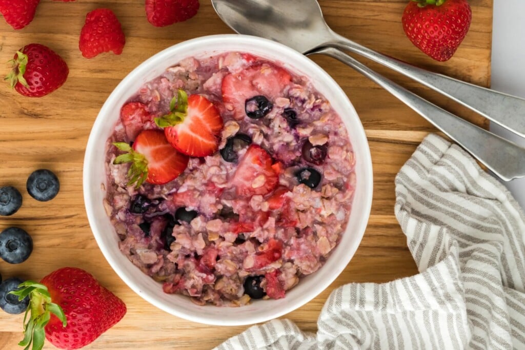 A bowl of oatmeal with chopped strawberries and blueberries laid out on a wooden board, surrounded by fresh berries, spoons and a striped napkin—the perfect inspiration for high-fiber breakfast recipes.