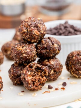 A stack of Chocolate Peanut Butter Energy Bites sits on a white marble board, with more bites and scattered oats around. A small bowl of mini chocolate chips and blurred ingredients are in the background.