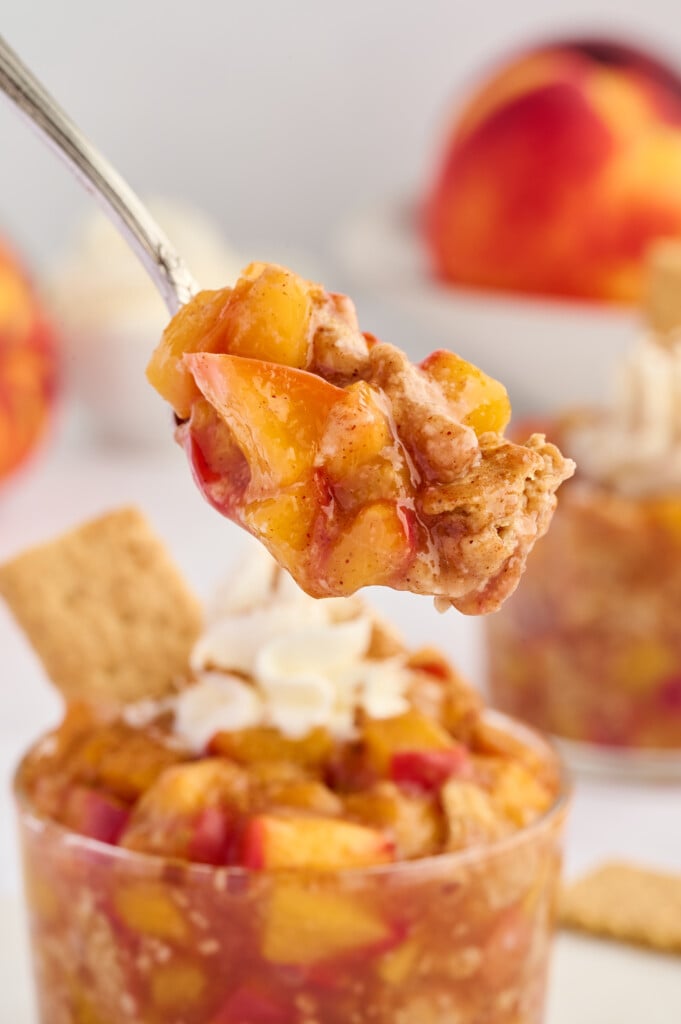 A spoon holding a bite of peach dessert with cinnamon and whipped cream, above a glass filled with the same Easy 5-Minute Peach Cobbler and a graham cracker. Blurry peaches and another dessert are in the background.