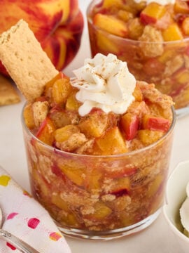 A glass cup filled with peach cobbler topped with whipped cream and a sprinkle of cinnamon, garnished with a graham cracker. Another dessert and a peach are in the background.