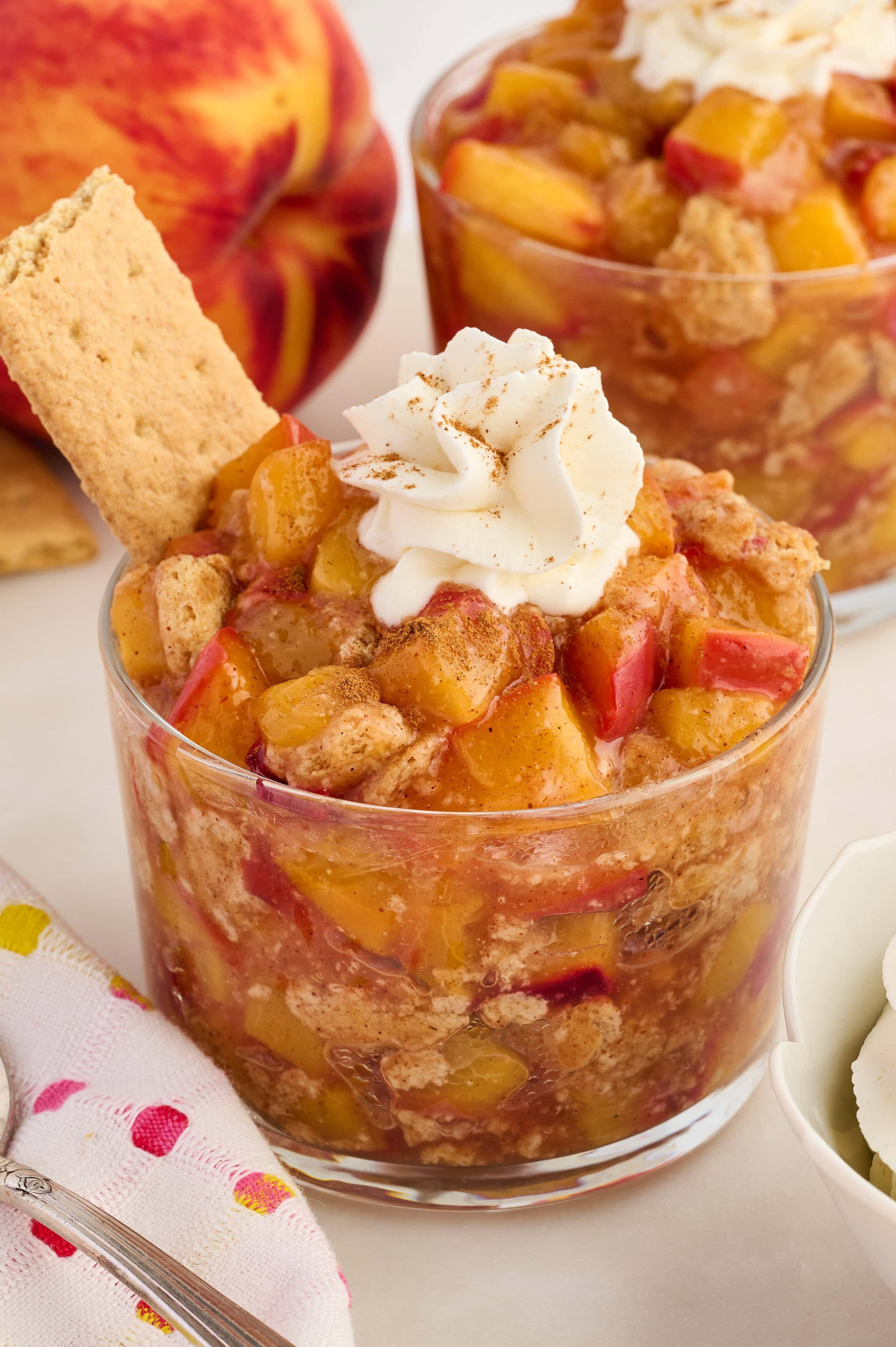 A glass cup filled with peach cobbler topped with whipped cream and a sprinkle of cinnamon, garnished with a graham cracker. Another dessert and a peach are in the background.