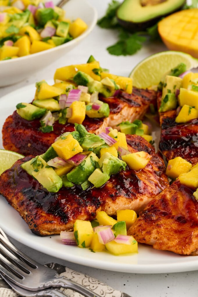 Grilled Barbecue Salmon Steaks with Avocado-Mango Salsa are beautifully presented on a white plate, topped with fresh mango avocado salsa, red onion, and cilantro. Served alongside lime wedges and a crisp salad in the background, this dish is as vibrant as it is flavorful.
