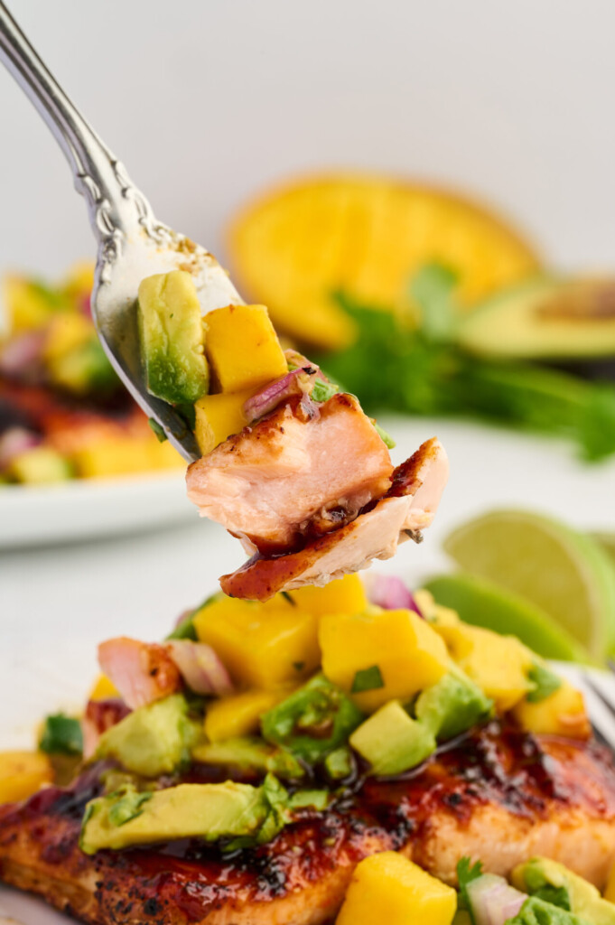 A fork holds a bite of grilled barbecue salmon steak topped with avocado-mango salsa and red onion, with more of the Grilled Barbecue Salmon Steaks with Avocado-Mango Salsa and fresh ingredients like lime and mango visible in the background.