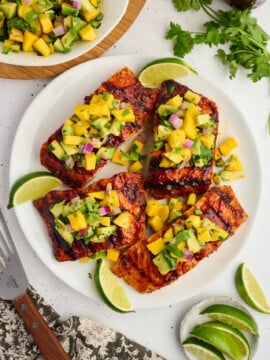 Four Grilled Barbecue Salmon Steaks with Avocado-Mango Salsa are served on a white plate alongside fresh lime wedges. Extra salsa and a bunch of cilantro are nearby, while a fork and a sliced lime complete the vibrant table setting.