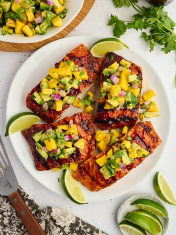 Four Grilled Barbecue Salmon Steaks with Avocado-Mango Salsa are served on a white plate alongside fresh lime wedges. Extra salsa and a bunch of cilantro are nearby, while a fork and a sliced lime complete the vibrant table setting.