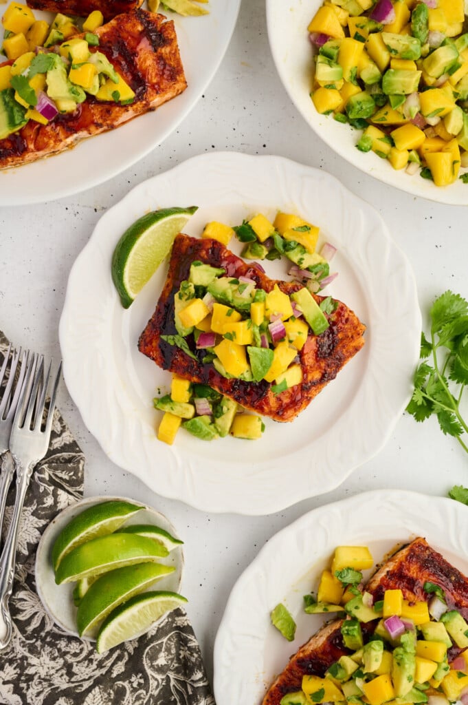 Two white plates feature Grilled Barbecue Salmon Steaks topped with vibrant Avocado-Mango Salsa, garnished with lime wedges. Extra salsa and fresh lime slices are served on the side, accompanied by forks and a brown napkin.